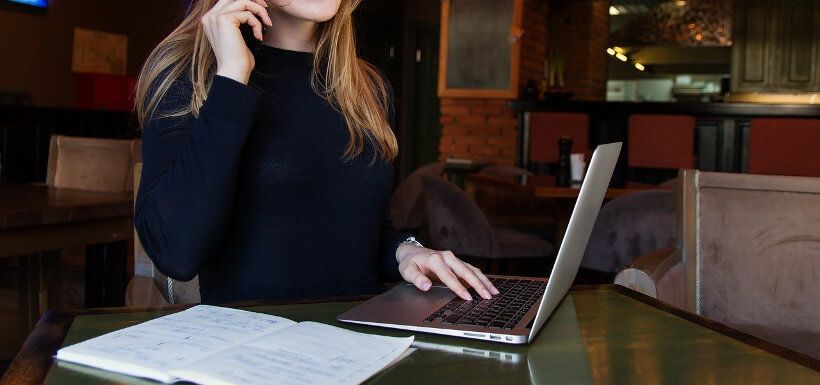 eine junge Frau, deren Gesicht nicht im Bildausschnitt ist, sitzt in einem Café vor Laptop und Notizbuch und scheint mit jemandem zu telefonieren