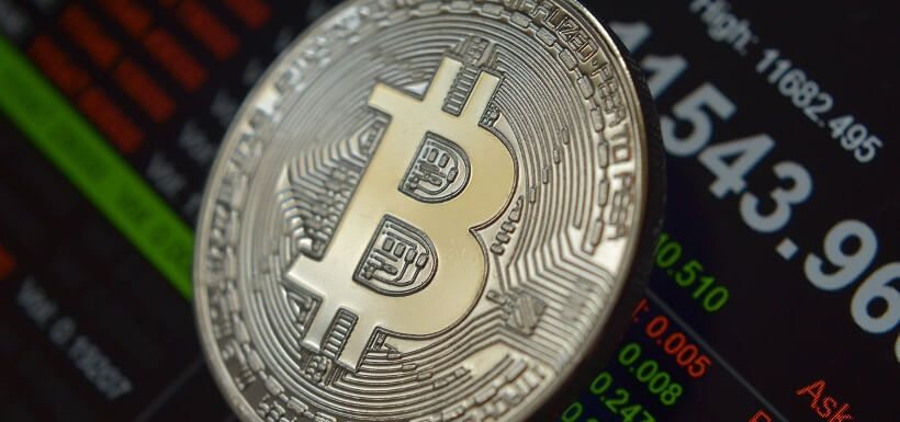 eine Bitcoin-Münze im Vordergrund, eine Börsenübersicht im Hintergrund