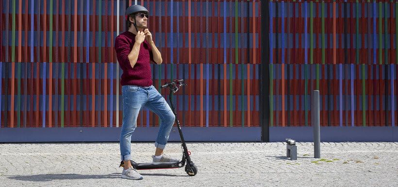 ein junger Mann steht lässig mit einem Fuß auf einem eScooter, während er sich einen Fahrrad-Helm an- bzw. auszieht