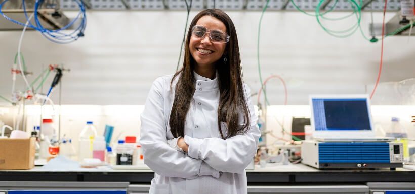 junge Frau mit Schutzbrille und weißem Kittel posiert vor einem chemischen Labor